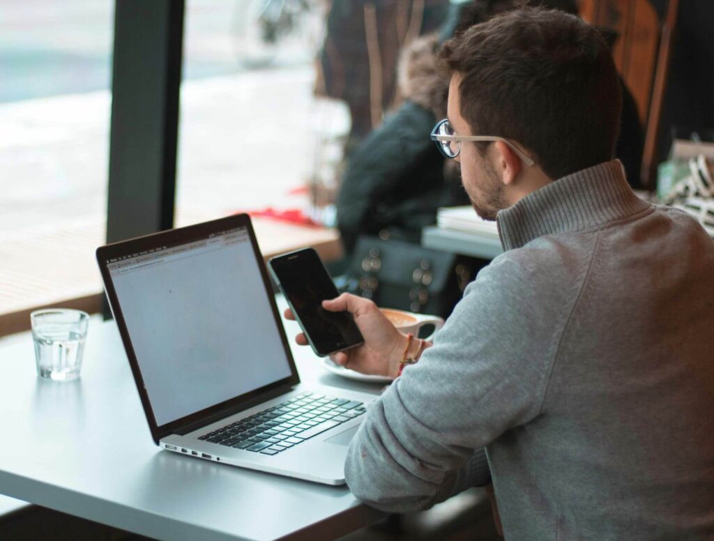 Man looking at his cell phone sitting in front of his laptop